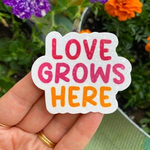 May include: A white sticker with the words "LOVE GROWS HERE" in pink and orange lettering. The sticker has a glittery effect and is held in front of a background of green plants and colorful flowers.
