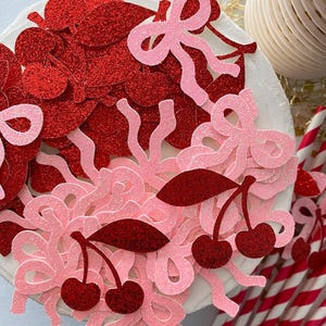 May include: Red and pink glitter confetti with cherry and bow shapes. The confetti is scattered on a white surface.