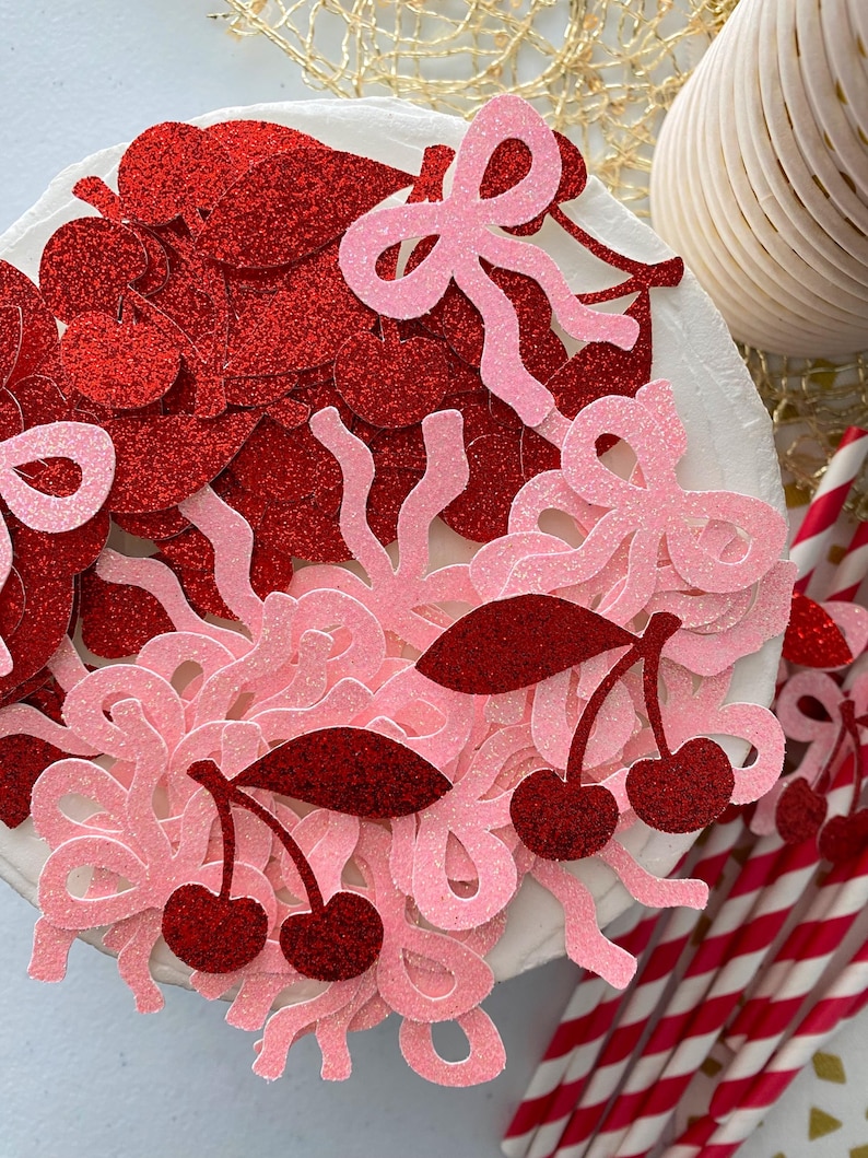 May include: Red and pink glitter confetti with cherry and bow shapes. The confetti is scattered on a white surface.