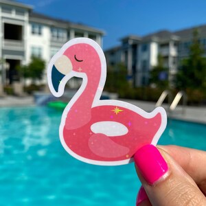 May include: A pink flamingo sticker with a dark blue beak and a sparkling design. The sticker is held in front of a pool of water and a blue sky.