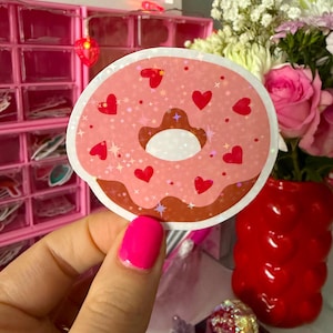 May include: A pink donut sticker with red heart and star accents. The donut has a brown frosting detail and a white center. The sticker is held in front of a pink background with a red vase of flowers.