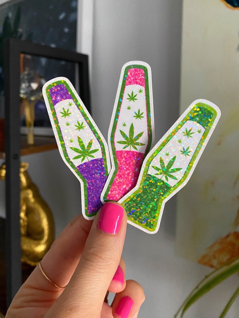 May include: Three lava lamp stickers with a green marijuana leaf design. The stickers are holographic and have a glitter finish. The stickers are purple, pink, and green.