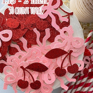 May include: A close-up of a white cake decorated with red and pink glitter confetti. The confetti is in the shape of cherries, bows, and swirls. The text "Great size! 1.75 - 2 inch 50 COUNT TOTAL" is visible on the cake.