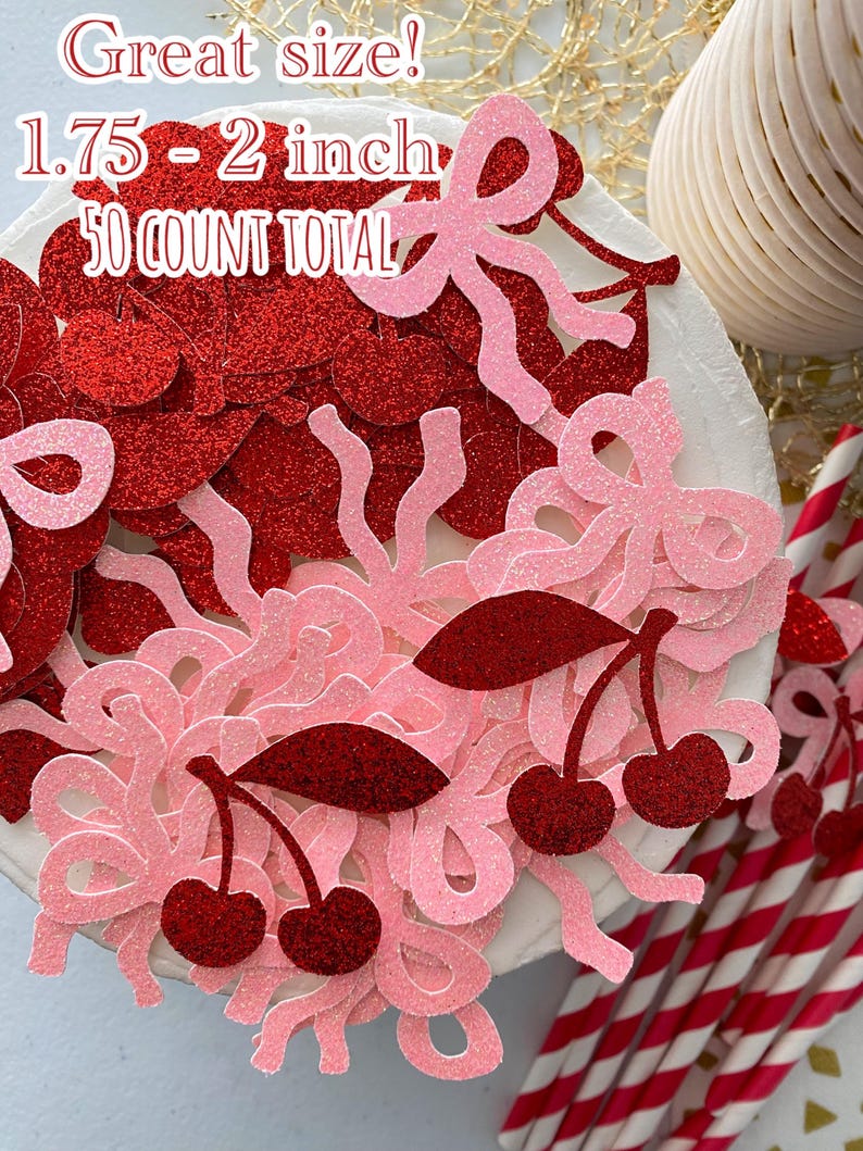 May include: A close-up of a white cake decorated with red and pink glitter confetti. The confetti is in the shape of cherries, bows, and swirls. The text "Great size! 1.75 - 2 inch 50 COUNT TOTAL" is visible on the cake.