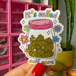 May include: A sticker featuring a cartoon jar filled with green objects, with the text "It's called self care." The jar has a pink lid and is surrounded by colorful flowers and stars. The sticker is held by a hand with red nail polish.
