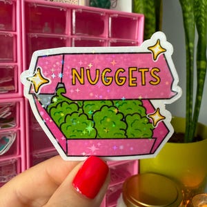 May include: A pink sticker with the word "NUGGETS" in yellow letters. The sticker depicts a box filled with green nuggets, with star accents and a holographic effect. The sticker is held by a hand with a red painted nail.