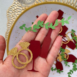May include: Gold glitter paper cutouts of a wine glass, wine bottle, and two rings. Green paper cutouts of vines are scattered around the cutouts.