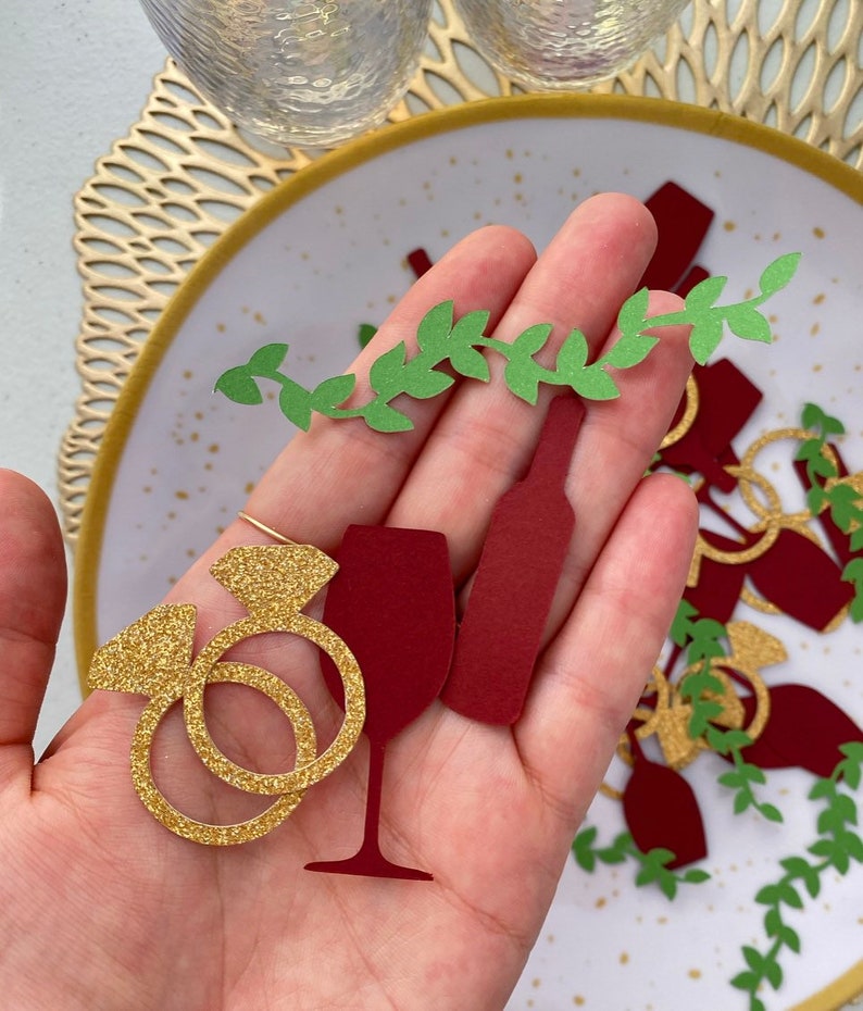 May include: Gold glitter paper cutouts of a wine glass, wine bottle, and two rings. Green paper cutouts of vines are scattered around the cutouts.