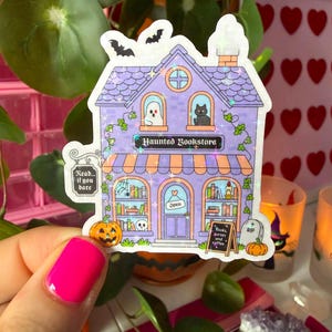 May include: A holographic sticker of a two-story purple building labeled Haunted Bookstore. The design features a ghost, a black cat, pumpkins, a tombstone, and a sign that reads Books, curses and coffee. Two bats fly above the roof.