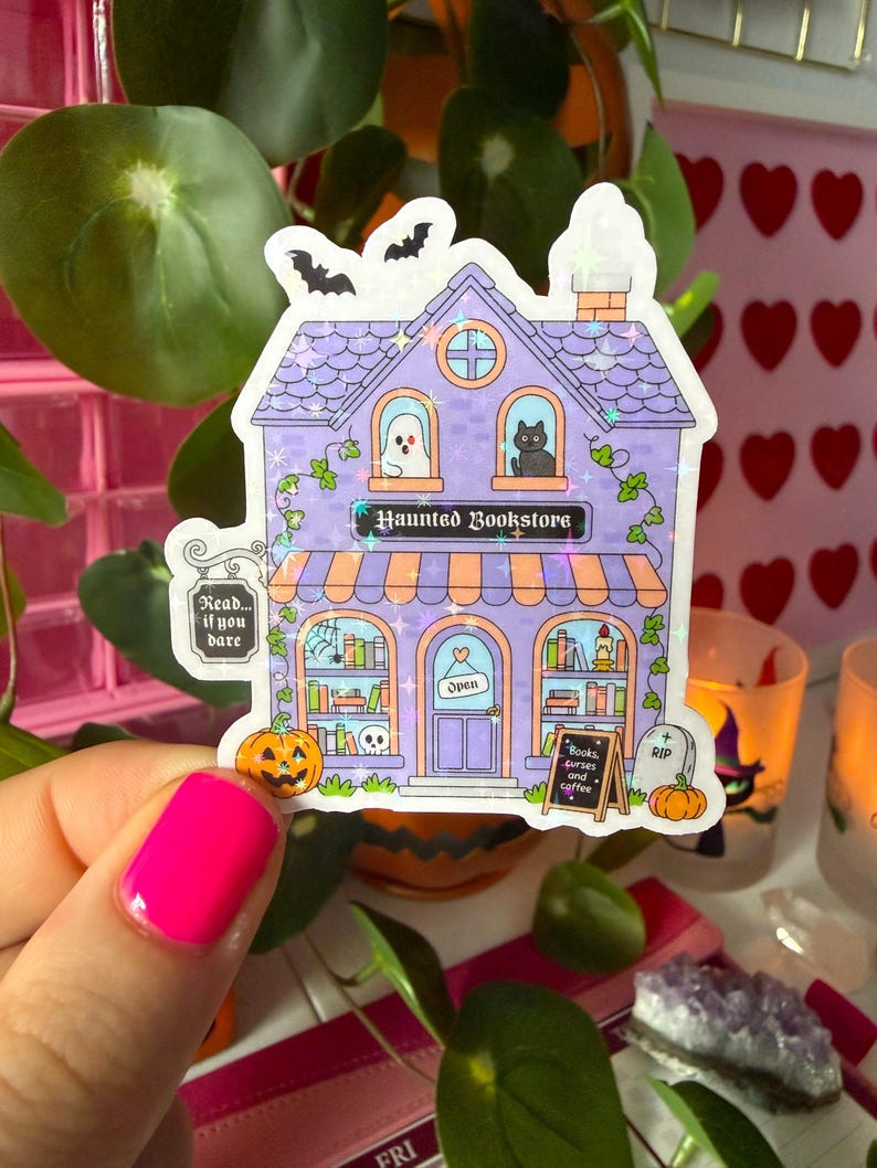 May include: A holographic sticker of a two-story purple building labeled Haunted Bookstore. The design features a ghost, a black cat, pumpkins, a tombstone, and a sign that reads Books, curses and coffee. Two bats fly above the roof.