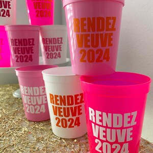Peut inclure: Un groupe de gobelets en plastique rose et blanc avec le texte "Rendez Vous 2024" imprim&eacute; en orange et blanc.