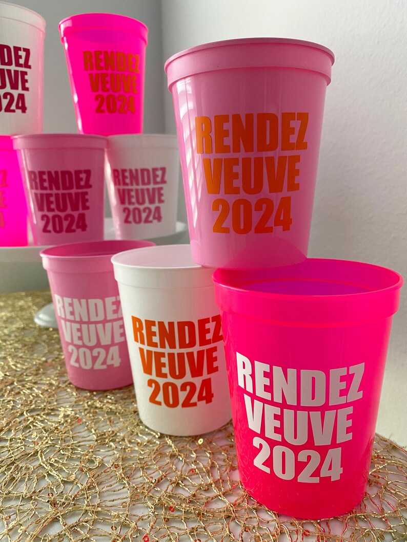 Peut inclure: Un groupe de gobelets en plastique rose et blanc avec le texte "Rendez Vous 2024" imprim&eacute; en orange et blanc.