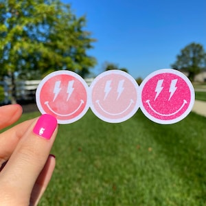 May include: Three pink and white smiley face stickers with lightning bolts. The stickers are in a gradient of pink, with the darkest pink at the bottom and the lightest pink at the top.