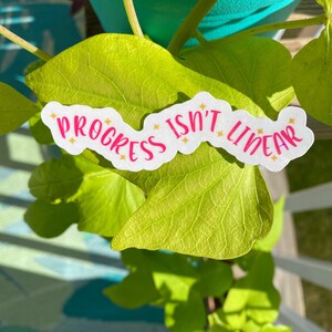 May include: A white sticker with pink lettering that reads "Progress isn't linear" with gold glitter accents. The sticker is placed on a green leaf.