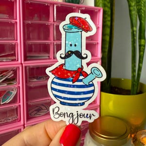 May include: A sticker featuring a cartoon-style bong with a red beret, mustache, and scarf. The bong has a blue and white striped design with the text "Bong jour" in a script font.