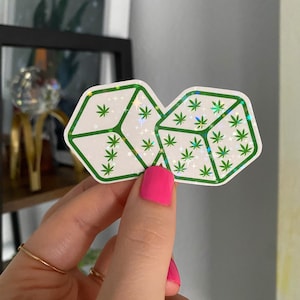 May include: A holographic sticker with a green marijuana leaf pattern. The sticker is shaped like two connected diamonds.
