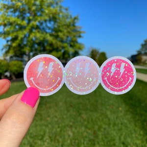 May include: Three pink and white smiley face stickers with a lightning bolt design. The stickers have a holographic glitter finish.