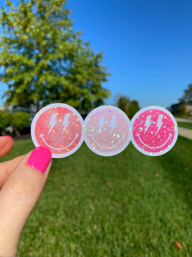 May include: Three pink and white smiley face stickers with a lightning bolt design. The stickers have a holographic glitter finish.