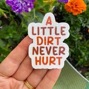 May include: A white sticker with the text "A LITTLE DIRT NEVER HURT" in orange and brown lettering. The sticker is held in front of a background of green foliage and colorful flowers.