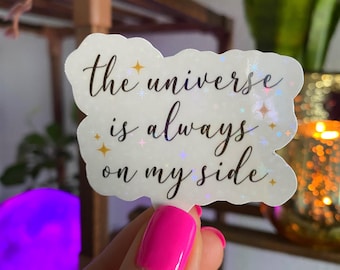 Op de afbeelding: Een witte, iriserende sticker met de tekst "the universe is always on my side" in zwarte letters. De sticker heeft een wolkvorm en is versierd met kleine gouden en zilveren glittersterren.