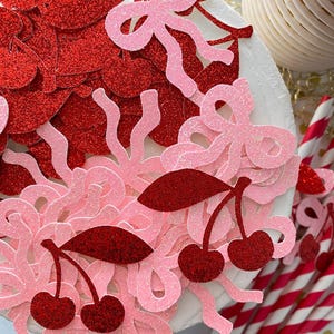 May include: Pink and red glitter confetti with cherry, bow, and noodle shapes. The confetti is scattered on a white surface.