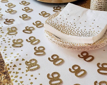 May include: A white table covered in a white tablecloth with gold confetti and gold glitter numbers 90 scattered across the surface. There is a white cake on a gold cake stand, a stack of gold confetti napkins, and a gold glitter party favor.
