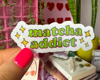 Op de afbeelding: Een holografische sticker met de woorden "matcha addict" in groene bubbelletters met een gele omlijning. De sticker is omgeven door gele sterren. De achtergrond bevat boeken, kristallen en een roze container.