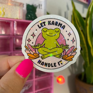 May include: A holographic sticker with a pink background and a green frog meditating with the text "Let Karma Handle It" in a white circle.