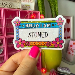 May include: A rectangular sticker with the text "HELLO I AM STONED" in a colorful floral frame. The sticker has a holographic background and is held by a hand with bright pink nail polish.