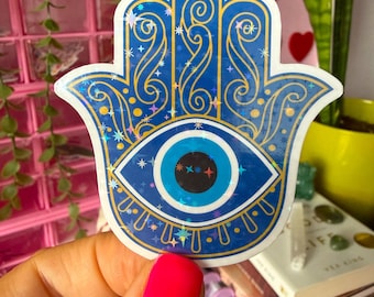 Op de afbeelding: Een blauwe en gouden Hamsa hand sticker met een evil eye design. De sticker heeft een gedetailleerd patroon met sterren en krullen. Het oog is helderblauw met een zwarte pupil en witte accenten. De sticker heeft een holografische, glitterende afwerking.