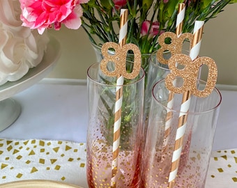 May include: Three clear glass cups with gold glitter accents and white and gold striped straws. Each straw has a gold glitter number "80" attached to it. The cups are on a white table with a gold confetti runner.