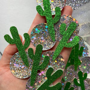May include: A set of green glitter cactus cutouts on a silver iridescent glitter background. The cactus shapes are simple and modern, with a single stem and two arms.