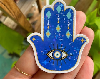 May include: A blue hamsa hand sticker with a gold outline and a blue evil eye in the center. The sticker is covered in glitter and has a white background.