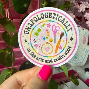 Może przedstawiać: Okrągła naklejka z holograficznym wykończeniem zawierająca napis UNAPOLOGETICALLY in the arts and crafts aisle. Wzór zawiera ilustracje tubki z farbą, palety, różowych nożyczek, pędzla oraz tamborka do haftowania.