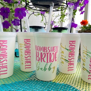 May include: White plastic cups with lids and straws. The cups have pink and green text that says "Bombshell" and names like "Gabby" and "Veronica". The cups are on a blue and green woven surface, with purple flowers in the background.