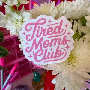 May include: A sticker with the text "Tired Moms Club" in pink script with glitter accents. The sticker is white with a cloud-like shape. The sticker is placed in a bouquet of white flowers.