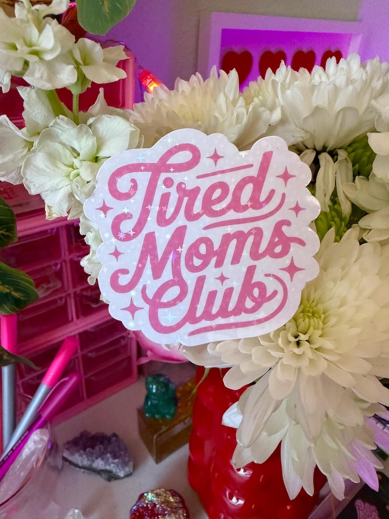 May include: A sticker with the text "Tired Moms Club" in pink script with glitter accents. The sticker is white with a cloud-like shape. The sticker is placed in a bouquet of white flowers.
