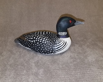 May include: A hand-carved wooden loon with black and white markings. The loon has a black head with a red eye and a long, thin beak.