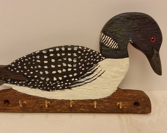 Puede incluir: Llavero de madera con forma de somorgujo. El ave está pintada con colores negro, blanco y marrón, con un ojo y un pico rojos. El llavero tiene cinco ganchos de latón.