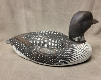 May include: A hand-carved wooden loon decoy with black and white paint. The decoy is lying on its side with its head turned to the right. The decoy is made of wood and has a realistic appearance.