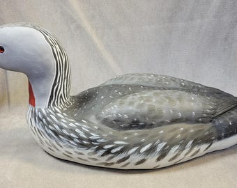 May include: Hand-carved wooden loon figurine with gray and white painted feathers, a red beak, and black eyes. The loon is lying on its side with its head turned to the left.
