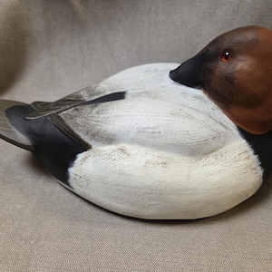 May include: A hand-carved wooden duck decoy with a white and gray body, black tail feathers, and a brown head with a red eye. The decoy is lying on its side on a brown fabric background.