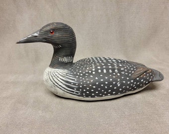May include: A hand-carved wooden loon figurine with black and white markings. The loon has red eyes and a long, thin beak.