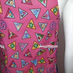 May include: Pink fabric apron with a white lace trim and a pocket. The apron is covered in a repeating pattern of brown triangles with various Girl Scout badges inside. The badges include a book, a musical note, a swirl, a bear, a giraffe, and a girl scout logo.
