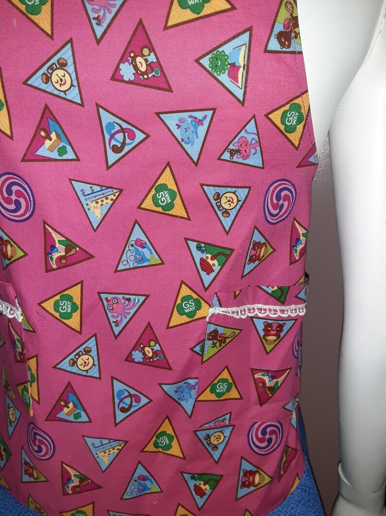 May include: Pink fabric apron with a white lace trim and a pocket. The apron is covered in a repeating pattern of brown triangles with various Girl Scout badges inside. The badges include a book, a musical note, a swirl, a bear, a giraffe, and a girl scout logo.