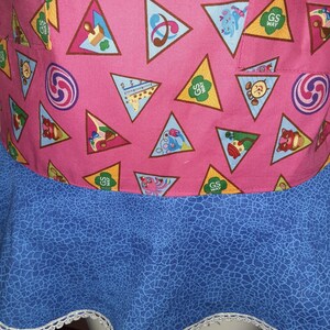 May include: A pink apron with a blue bottom and white lace trim. The apron is covered in brown triangles with various Girl Scout badges inside. The badges include a musical note, a compass, a giraffe, a squirrel, a butterfly, and a clock.