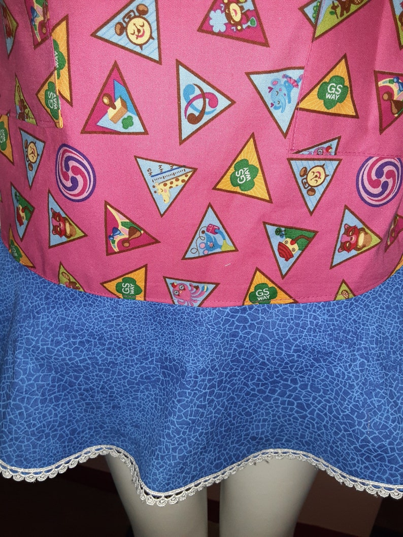 May include: A pink apron with a blue bottom and white lace trim. The apron is covered in brown triangles with various Girl Scout badges inside. The badges include a musical note, a compass, a giraffe, a squirrel, a butterfly, and a clock.