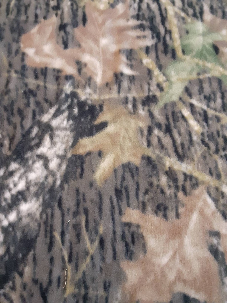May include: Camouflage fabric with a brown, green and black pattern. The fabric has a soft, fuzzy texture.