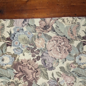 May include: A floral tapestry table runner featuring a cream background with large, detailed flowers in shades of mauve, blue, and green. The runner is placed on a dark wooden surface, showcasing the intricate floral design.