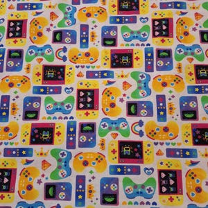 Könnte beinhalten: Ein weißer Stoff mit einem sich wiederholenden Muster aus bunten Videospiel-Controllern, Handheld-Konsolen und anderen Gaming-Symbolen. Das Design umfasst gelbe, blaue, grüne und violette Controller sowie Sterne, Herzen und Kronen.